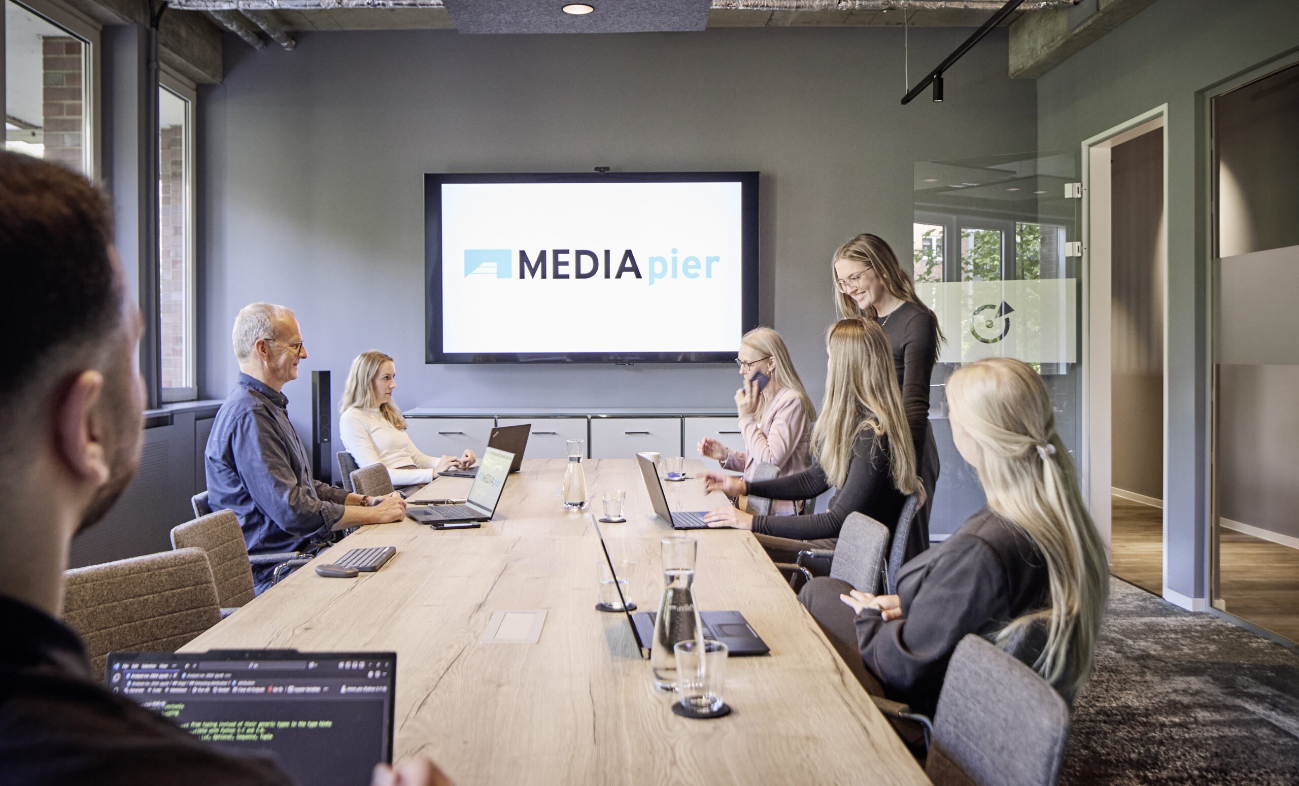 Sechs Personen versammeln sich in einem modernen Konferenzraum, einige mit Laptops, um einen langen Tisch. Einer steht und präsentiert neben einem Bildschirm mit dem Logo "MEDIA pier" - eine Full-Service-Medienagentur. Große Fenster füllen den Büroraum mit Licht.